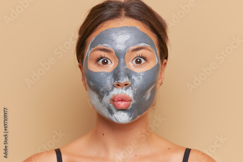 Portrait of young woman with facial mask made from clay posing over studio background, skincare concept, copy space