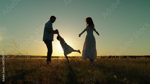 Family enjoys tranquility of park among waving grass forming memories on journey. Mother dances and father spins daughter holding hands at dusk. Family silhouettes in field of swaying grass at sunset