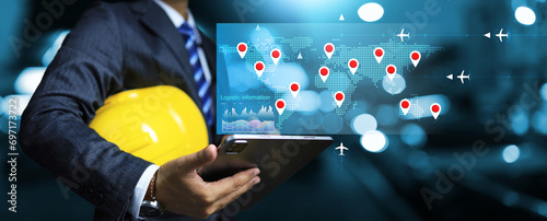 Logistic concept with businessman holding yellow helmet and monitoring tracking industrial cargo shipment on world map flight transportation information to confirm order and prepare production plan