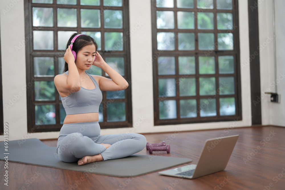 A young Asian woman with headphones practicing Yoga online with a trainer learning laptop on a yoga mat at home.