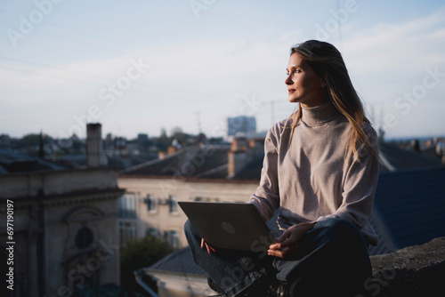 Woman freelancer uses laptop on cement wall outdoors against the sky and the roof of the city. The woman to be focused on her work or enjoying some leisure time while using her laptop.