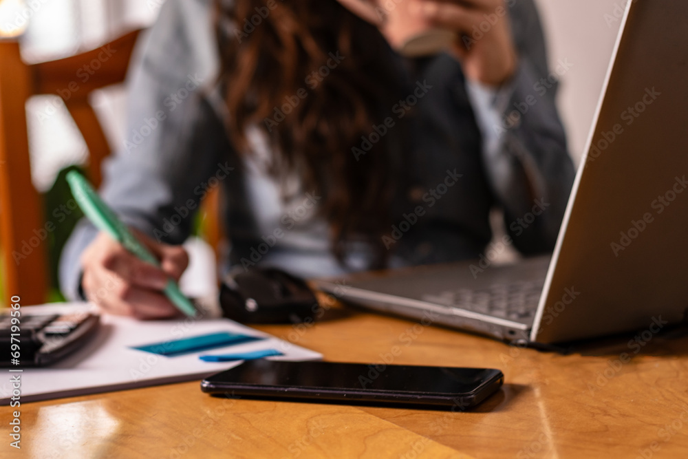 small businesswoman is sitting at her desk at home, drinking her coffee, checking her bank and making notes. On the table you can see her smart phone. small businesswoman is sitting at her desk at home, drinking her coffee, checking her bank and making notes. On the table you can see her smart phone.