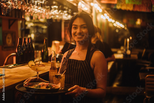 Portrait of smiling waitress holding food and drinks on serving tray while working at bar photo