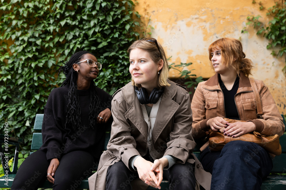 Young woman looking away while sitting with female friends on bench at park Young woman looking away while sitting with female friends on bench at park
