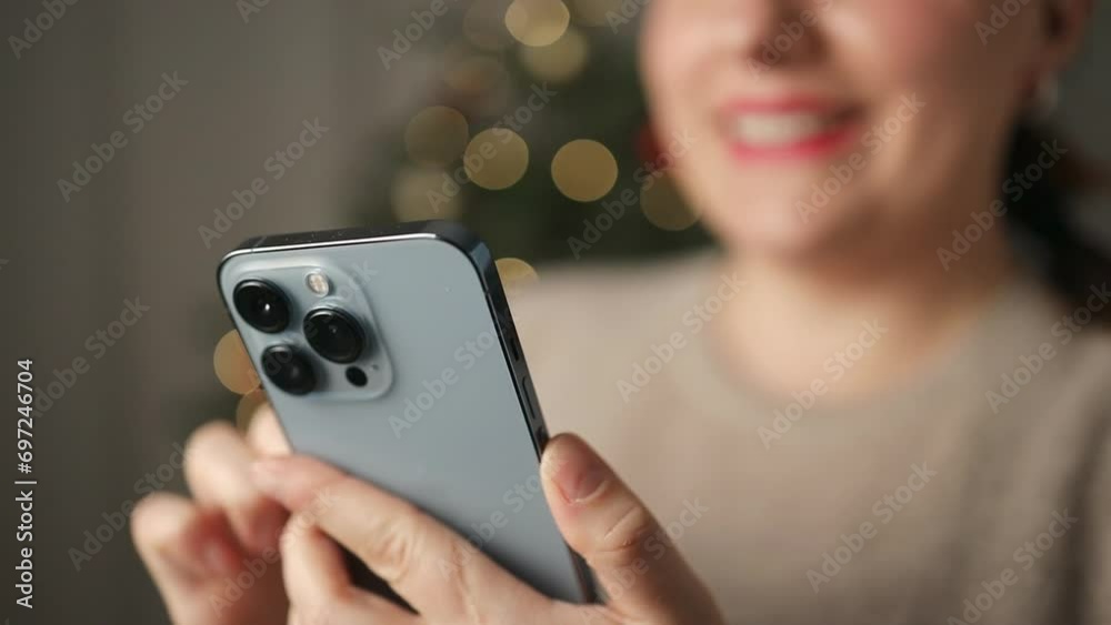 Smiling young woman uses a smartphone sitting at home during winter New Year Christmas Xmas holidays. Girl is writing a greeting message, chatting with friends or shopping online. High quality FullHD Smiling young woman uses a smartphone sitting at home during winter New Year Christmas Xmas holidays. Girl is writing a greeting message, chatting with friends or shopping online. High quality FullHD