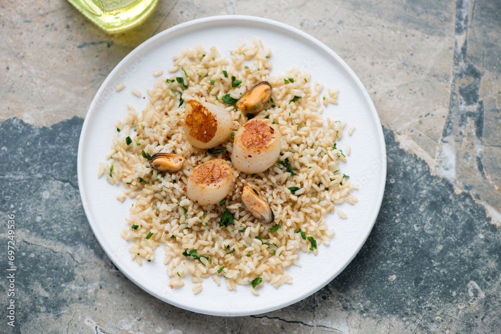 Seared scallops with brown rice and mussels on a white plate, horizontal shot on a grey and beige granite background, elevated view Seared scallops with brown rice and mussels on a white plate, horizontal shot on a grey and beige granite background, elevated view