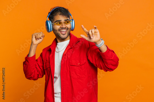 Happy stylish Indian man in sunglasses listening music via headphones, dancing disco fooling around having fun expressive gesticulating hands. Arabian excited guy isolated on orange background indoors photo