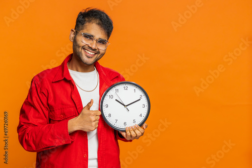 It is your time, hurry up. Indian young man showing time on wall office clock, ok, thumb up approve pointing finger at camera, advertisement, deadline. Arabian guy on orange background. Copy-space photo