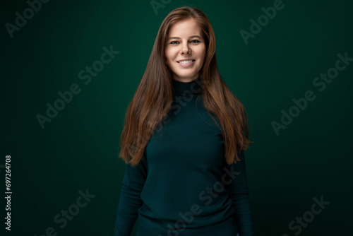 Cute modest woman with brown natural hair posing shyly on dark green background with copy space