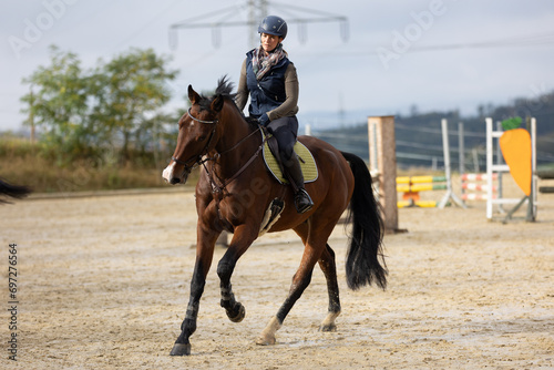 Horse during training with rider on the riding arena, galloping into a turn, photographed from the front with obstacles in the background.