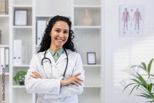 Portrait of young beautiful successful female doctor inside medical office, hispanic woman with curly hair in white medical coat smiling and looking at camera with arms crossed, doctor inside clinic.