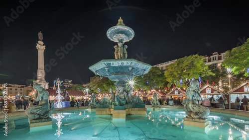 Panorama showing illuminated fountain with evening timelapse view of holiday decorations and food booths at the Rossio Christmas Market on Dom Pedro IV square in Baixa district of Lisbon, Portugal.