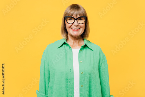 Elderly smiling happy cheerful fun satisfied blonde woman 50s years old wear green shirt glasses casual clothes looking camera isolated on plain yellow background studio portrait. Lifestyle concept.