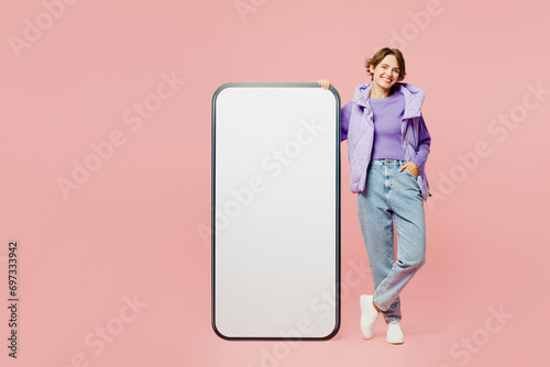Full body young fun woman wear purple vest sweatshirt casual clothes look camera stand near big huge blank screen mobile cell phone smartphone with area isolated on plain pastel light pink background.