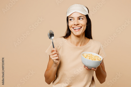 Young Latin woman wears pyjamas jam sleep eye mask rest relax at home eat breakfast muesli cereals with milk fruit in bowl look aside isolated on plain beige background. Good mood night nap concept.