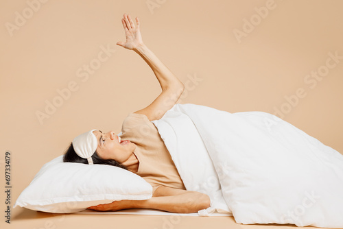 Outraged young sad mad Latin woman wears pyjamas jam sleep eye mask try to rest relax at home lay down under duvet scream shout isolated on plain pastel beige background. Bad mood night nap concept. photo