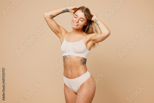 Young nice lady woman with slim body perfect skin wear nude top bra lingerie stand with raise up hands touch hair close eyes isolated on plain pastel light beige background Lifestyle diet fit concept