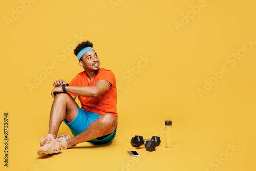 Full body fun young fitness trainer sporty man sportsman wear orange t-shirt use smart watch to check calories training in home gym isolated on plain yellow background. Workout sport fit abs concept.