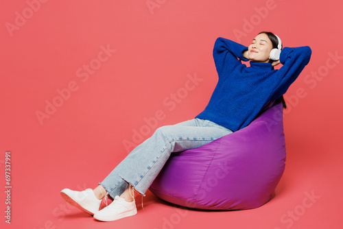 Full body serene young woman of Asian ethnicity she wear blue sweater casual clothes sit in bag chair rest listen to music in headphones isolated on plain pastel light pink background studio portrait.