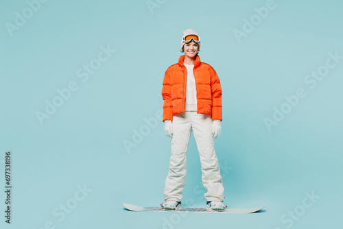 Full body young happy woman wearing warm padded windbreaker jacket hat ski goggles mask standing with snowboard travel rest spend weekend winter season in mountains isolated on plain blue background.