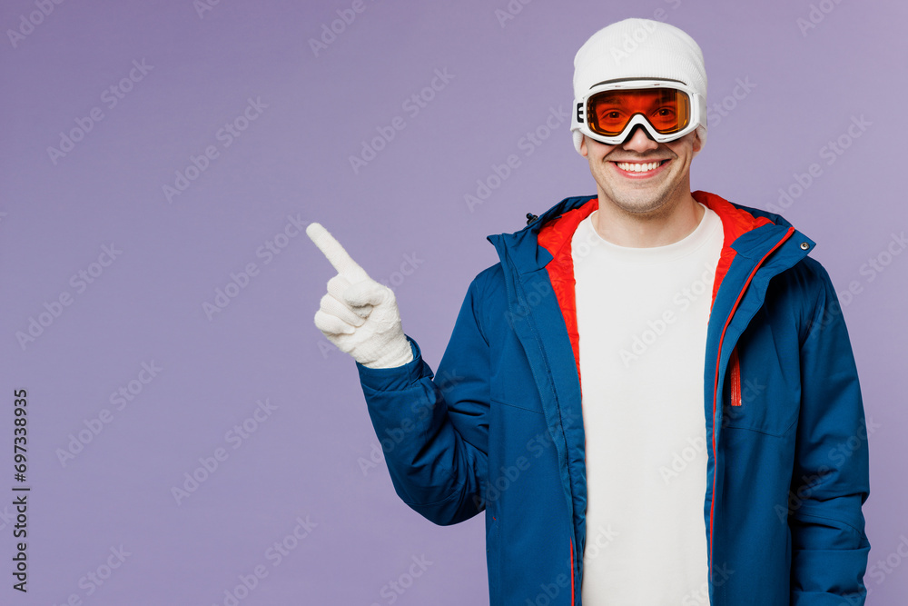Skier smiling happy man wears warm blue windbreaker jacket ski goggles mask hat point index finger aside on area spend extreme weekend winter season in mountains isolated on plain purple background. Skier smiling happy man wears warm blue windbreaker jacket ski goggles mask hat point index finger aside on area spend extreme weekend winter season in mountains isolated on plain purple background.