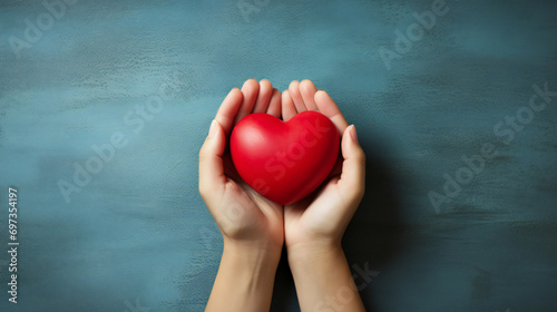 A person's hands delicately cradle a vibrant red heart, their fingertips adorned with colorful nails, as they hold it close to their chest in an intimate indoor setting