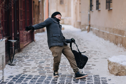 A young guy in a gray coat, hat and glove with a backpack walks along an old slippery snow-covered narrow street in Lviv, trying to resist and slipping. Christmas Eve, Ukraine.