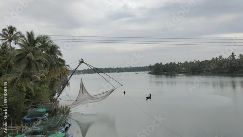 Aerial drone view of beautiful backwater lake landscape in Kerala with traditional Chinese fishing net and local fisherman rowing small wooden fishing boat surrounded with tropical coconut palm trees.