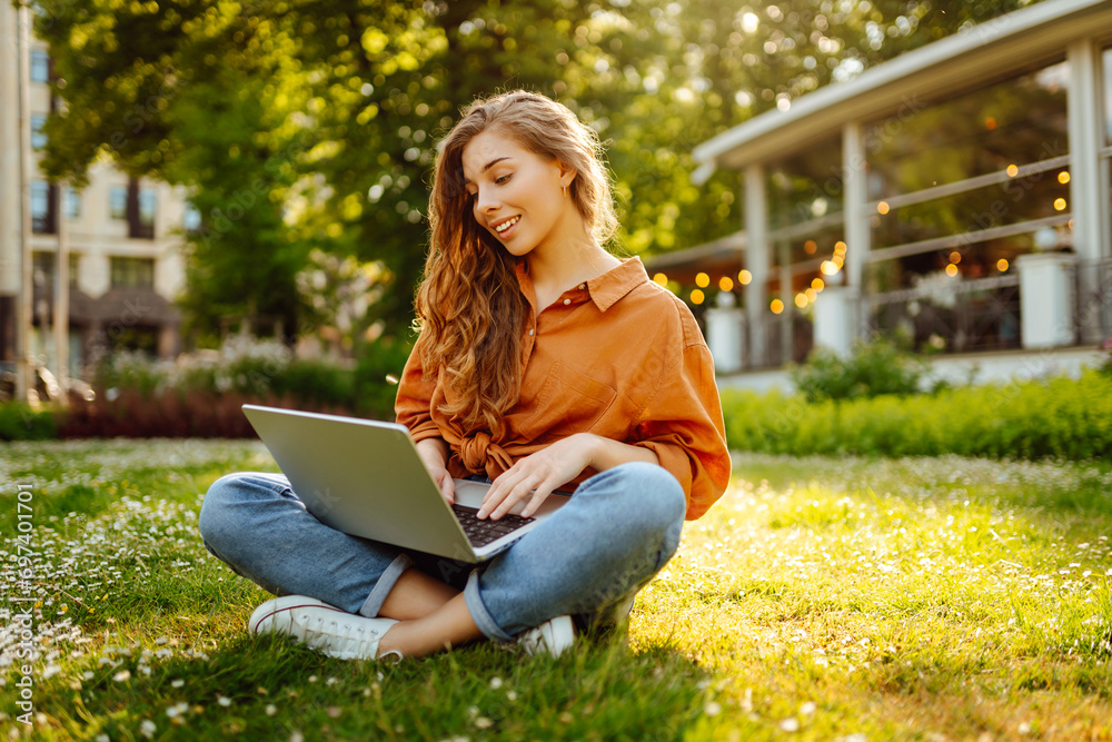 Portrait of a happy woman in a sunny park on a green lawn with a laptop. Young freelancer woman working on a laptop, enjoying the sunny weather. Freelance concept, technology. Portrait of a happy woman in a sunny park on a green lawn with a laptop. Young freelancer woman working on a laptop, enjoying the sunny weather. Freelance concept, technology.