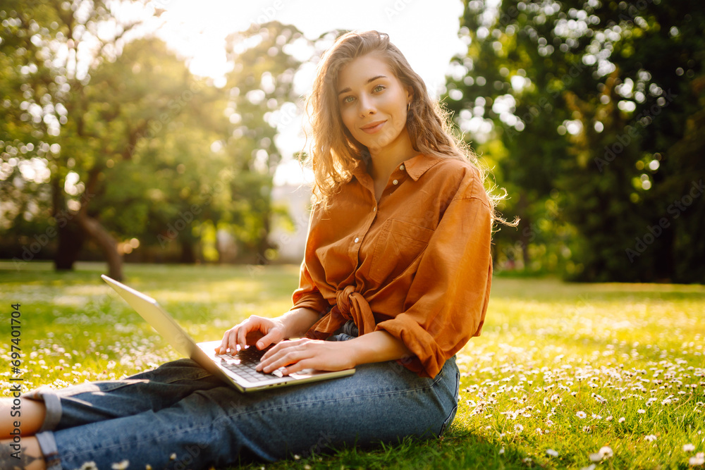 Portrait of a happy woman in a sunny park on a green lawn with a laptop. Young freelancer woman working on a laptop, enjoying the sunny weather. Freelance concept, technology. Portrait of a happy woman in a sunny park on a green lawn with a laptop. Young freelancer woman working on a laptop, enjoying the sunny weather. Freelance concept, technology.