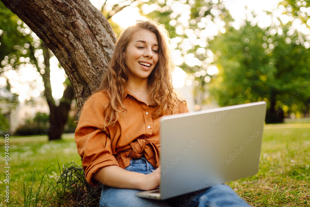Portrait of a happy woman in a sunny park on a green lawn with a laptop. Young freelancer woman working on a laptop, enjoying the sunny weather. Freelance concept, technology. Portrait of a happy woman in a sunny park on a green lawn with a laptop. Young freelancer woman working on a laptop, enjoying the sunny weather. Freelance concept, technology.