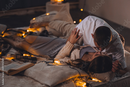 Beautiful happy loving young smiling couple relaxing in bed, watching movies on projector. Cozy home atmosphere, tenderness, closeness. Romantic surprise, atmospheric candles, Saint Valentine's Day photo
