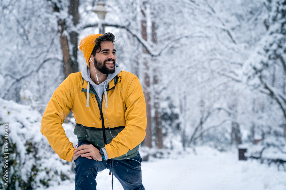 Handsome male runner warming up before running in city park while listening to music on snowy morning.