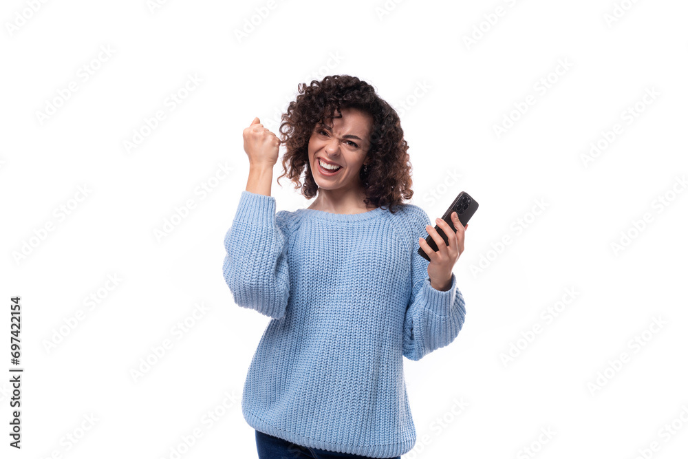 young caucasian woman with curly dark hair dressed in a casual warm blue jacket holds a smartphone in her hand rejoices young caucasian woman with curly dark hair dressed in a casual warm blue jacket holds a smartphone in her hand rejoices