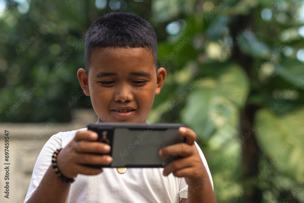 A young asian boy playing games on a large cellphone. Addictive leisure time for a kid. Outdoor garden scene. A young asian boy playing games on a large cellphone. Addictive leisure time for a kid. Outdoor garden scene.
