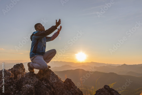Hiker male kneeling down with hands open palm up praying to God on top mountain sunset background. photo