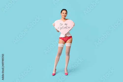 Beautiful young happy Asian woman in sexy underwear with paper heart on blue background. Valentine's Day celebration