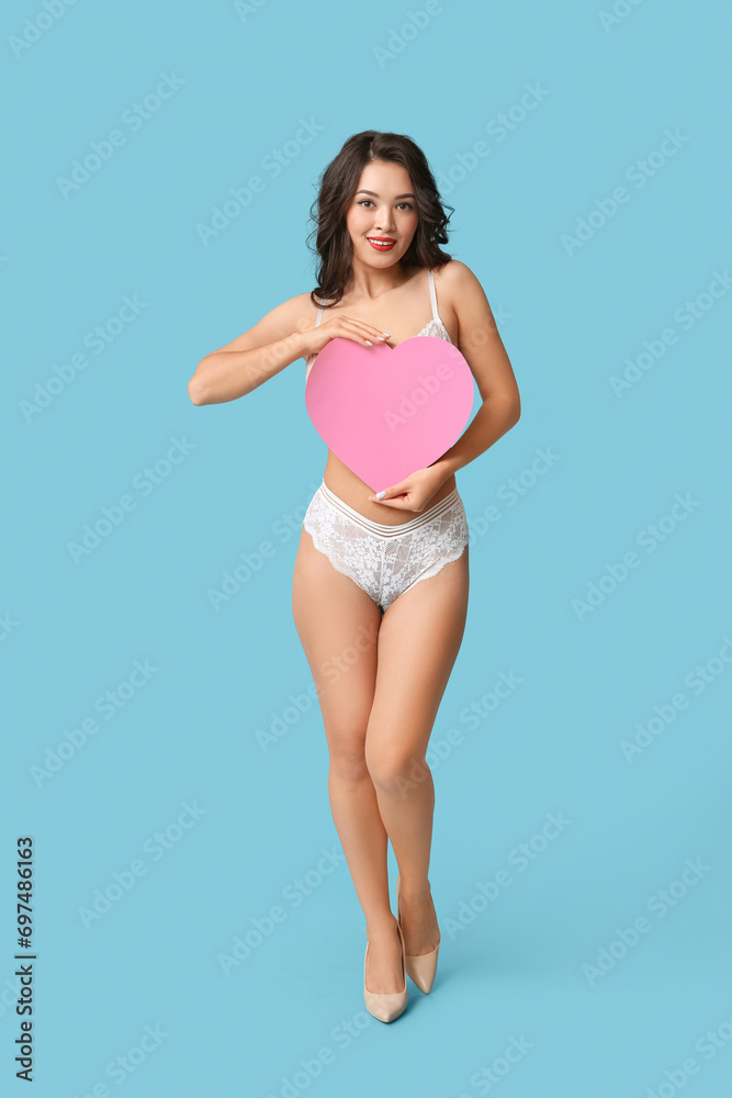 Naklejka premium Beautiful young happy Asian woman in sexy underwear with paper heart on blue background
