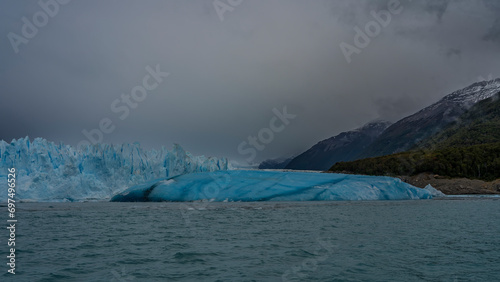 The beautiful glacier of Patagonia. A wall of cracked blue ice in a turquoise lake. A large breakaway iceberg floats in the water. Coastal mountains in clouds and fog. Perito Moreno. El Calafate. 