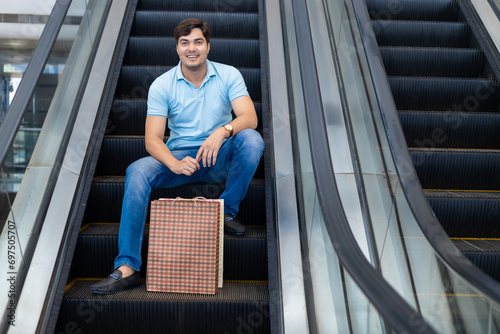 Happy young indian man sitting, riding escalator in shopping mall with paper bags, purchases. Shopaholic concept.
