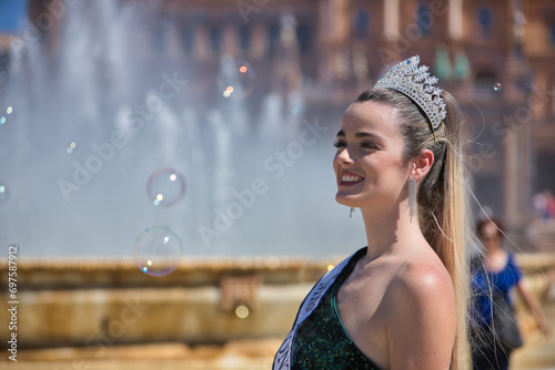 Foto Portrait of young, pretty, blonde woman in green sequined party outfit, with diamond crown and beauty pageant winner's sash, posing next to a fountain surrounded by soap bubbles