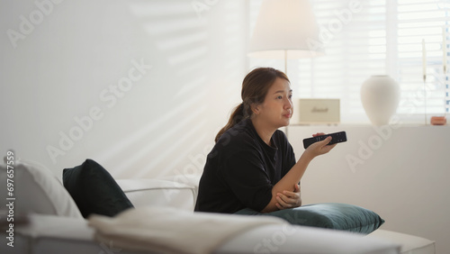 Woman sit down on sofa in living room at home searching for film or TV series to watch for leisure. She is using remote control choosing movie on streaming service.