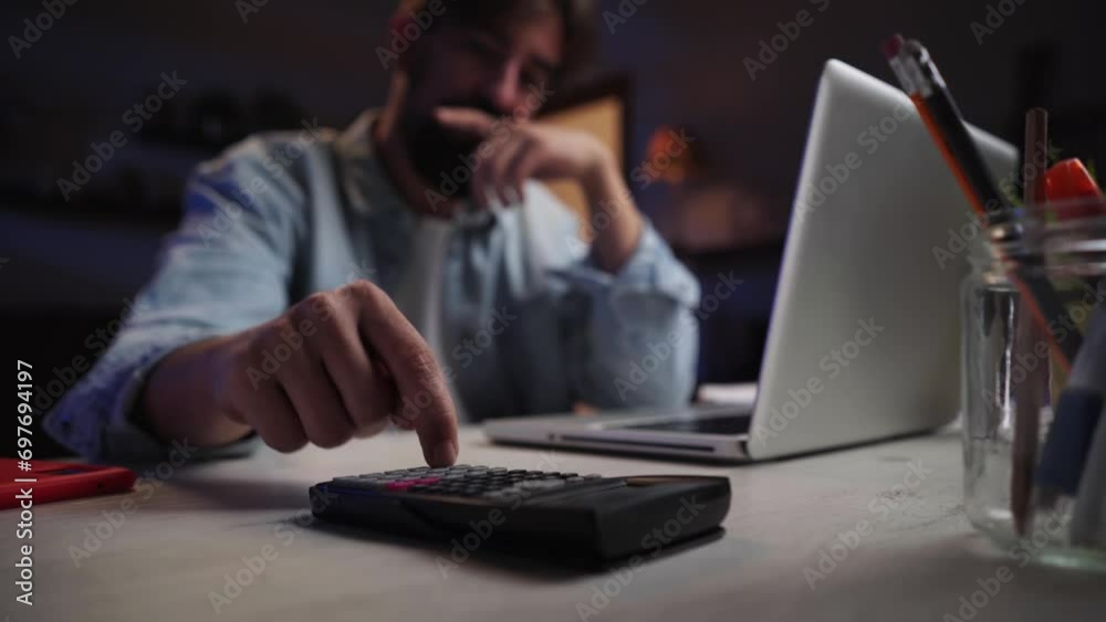 Young caucasian man using calculator to check his house finance and accounting, using a laptop late at home. Focus on calculator.