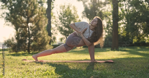 Yoga, woman gets into asana parshvakonasana pose, bent knee, palms together, balance. Side view, city park, lawn, green trees, morning sunlight. photo