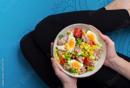 A woman in a sitting position on a blue fitness mat holds in her hands a bowl of salad with tuna and vegetables. Healthy nutrition concept.