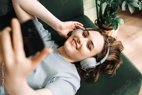 A smiling young middle-aged woman wearing wireless headphones listens to music while lying on the couch with her smartphone. Contentment in Tunes. Smiling Woman Enjoys Music with Wireless Headphones.
