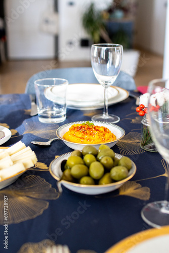 Dining table with water glasses, a bowl of hummus, and olives, decorated with cotton branches on a blue and gold cloth. Christmas table setup with olive and mediterranean food.