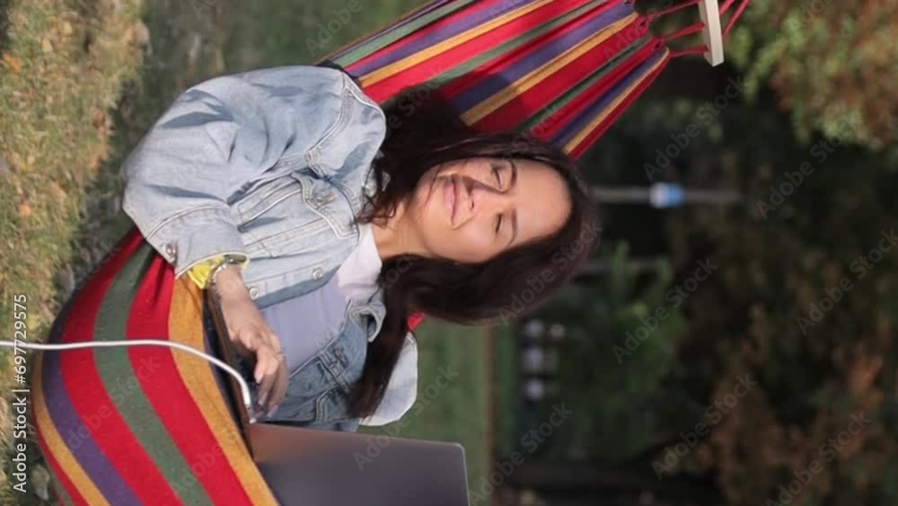 The girl is working on a laptop that is charging from a portal charging station. Resting on a hammock in the park. Vertical video The girl is working on a laptop that is charging from a portal charging station. Resting on a hammock in the park. Vertical video