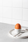Boiled egg in an egg stand and spoon on white tile kitchen background with text space. Light protein breakfast concept, minimalism, high key