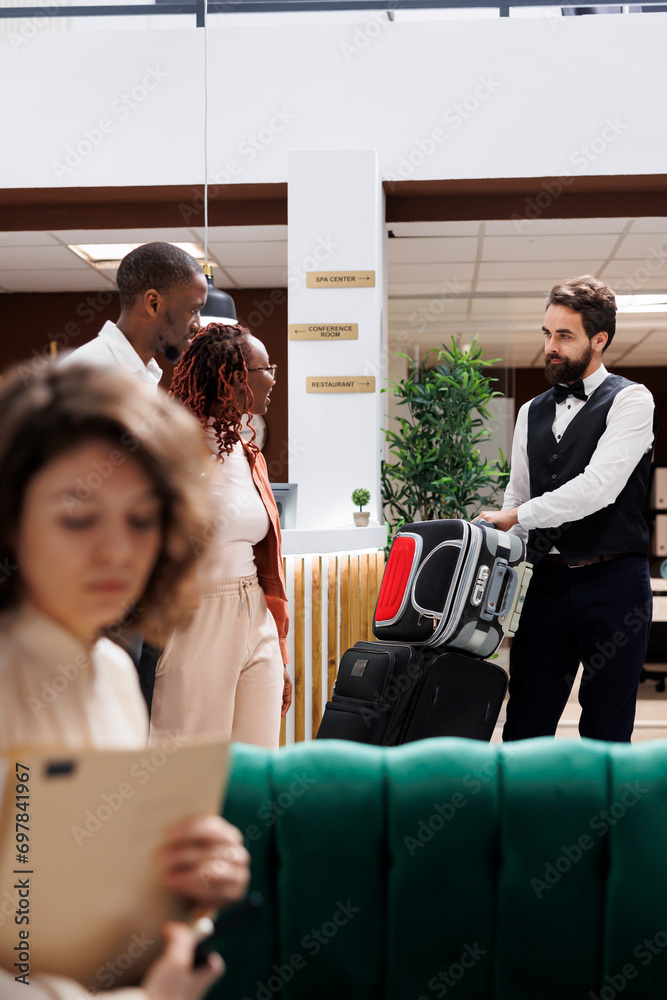 Male employee helping clients with bags, carrying luggage for people after doing check in procedure. Young couple receiving assistance from luxury resort bellboy at reception desk. Male employee helping clients with bags, carrying luggage for people after doing check in procedure. Young couple receiving assistance from luxury resort bellboy at reception desk.